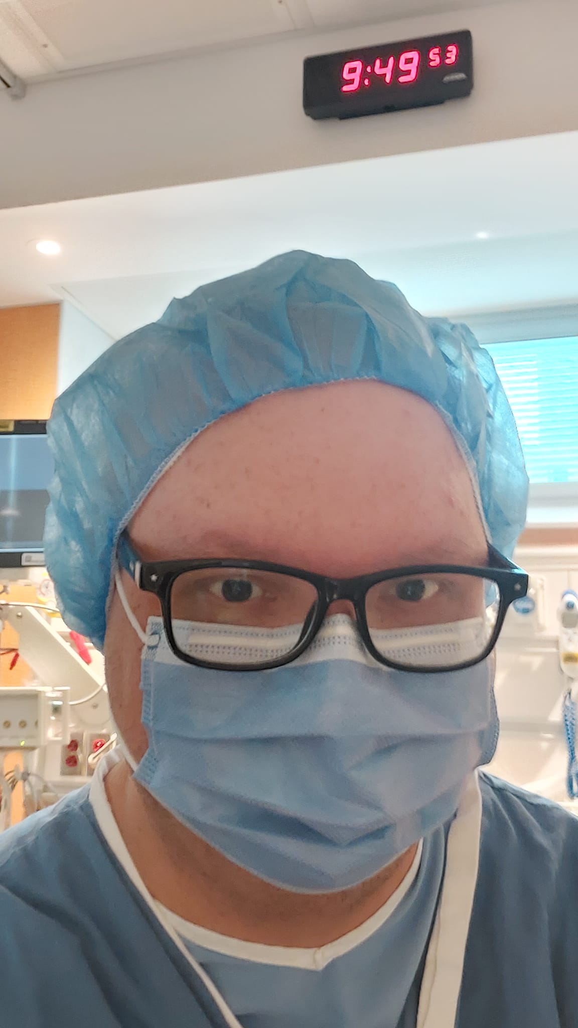 A man wearing a a surgical mask, blue surgical head covering and hospital robes sits upright on a hospital bed